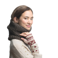 A person poses against a white background wearing the Dreams Hand Crochet Handwarmers—red-and-cream handwarmers handmade in Nepal—paired with a thick brown knitted scarf and a cream sweater with gold details.