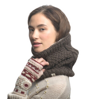 A woman with short brown hair wears a chunky dark brown knit scarf and the Dreams Hand Crochet Handwarmers, which feature patterns inspired by handmade Nepalese designs. She is dressed in a light sweater adorned with small pearl-like details and stands against a plain white background.