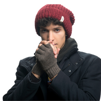 A young man wearing a red knit beanie, a dark coat, and the Hand Loom Striped and Solid Fingerless Gloves stands against a white background, holding his hands together near his face as if to warm them.