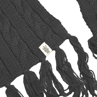 A close-up of a dark Hand Knit Plush Scarf, featuring thick cables, long fringe ends along the edge, and a small fabric tag with an intricate line design. Only sections of this Merino wool accessory are visible against a plain white background.