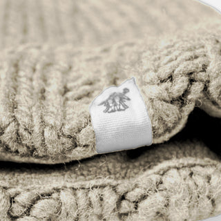 A close-up view of the Hand Knit Timber Hat in beige highlights its intricate wool texture, with a white tag featuring a small animal logo—possibly a horse—attached to the edge of the garment.