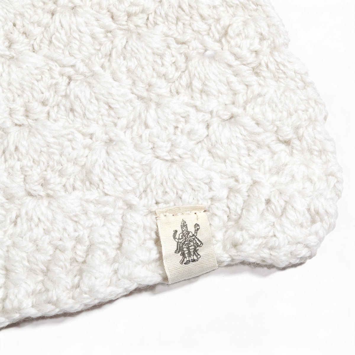 A close-up of the Hand Crochet Veil Scarf, crafted from white Merino wool in a textured bobble stitch. Near the edge, a small sewn-in label displays a black line drawing of a multi-armed figure.