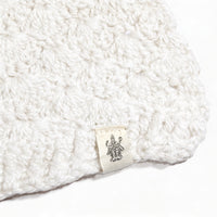 A close-up of the Hand Crochet Veil Scarf, crafted from white Merino wool in a textured bobble stitch. Near the edge, a small sewn-in label displays a black line drawing of a multi-armed figure.