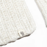 A close-up of the Hand Crochet Velour Scarf showcases its white knitted fabric with a distinctly woven texture, echoing a hand-crocheted or ribbed crochet pattern. A small tag with a printed black emblem or logo is visible against the plain white background.