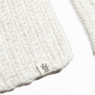 A close-up of the Hand Crochet Velour Scarf showcases its white knitted fabric with a distinctly woven texture, echoing a hand-crocheted or ribbed crochet pattern. A small tag with a printed black emblem or logo is visible against the plain white background.