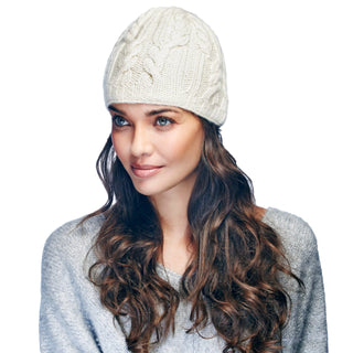 A woman with long, wavy hair wearing a Borderline Cable Knit Beanie handmade in Nepal and a light grey sweater, gazing to the side with a slight smile.