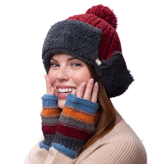 A smiling woman wearing a multicolored winter hat with ear flaps and a pom-pom, along with short cuff handwarmers.