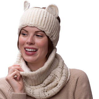 A woman in a beige sweater smiles slightly off-camera, wearing a white knitted hat with ears and a Lacey neckwarmer.