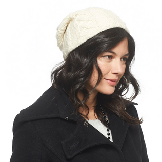 A woman with dark hair wearing a white Oslo Braid Knit Slouch handmade in Nepal merino wool slouch hat and a black coat.